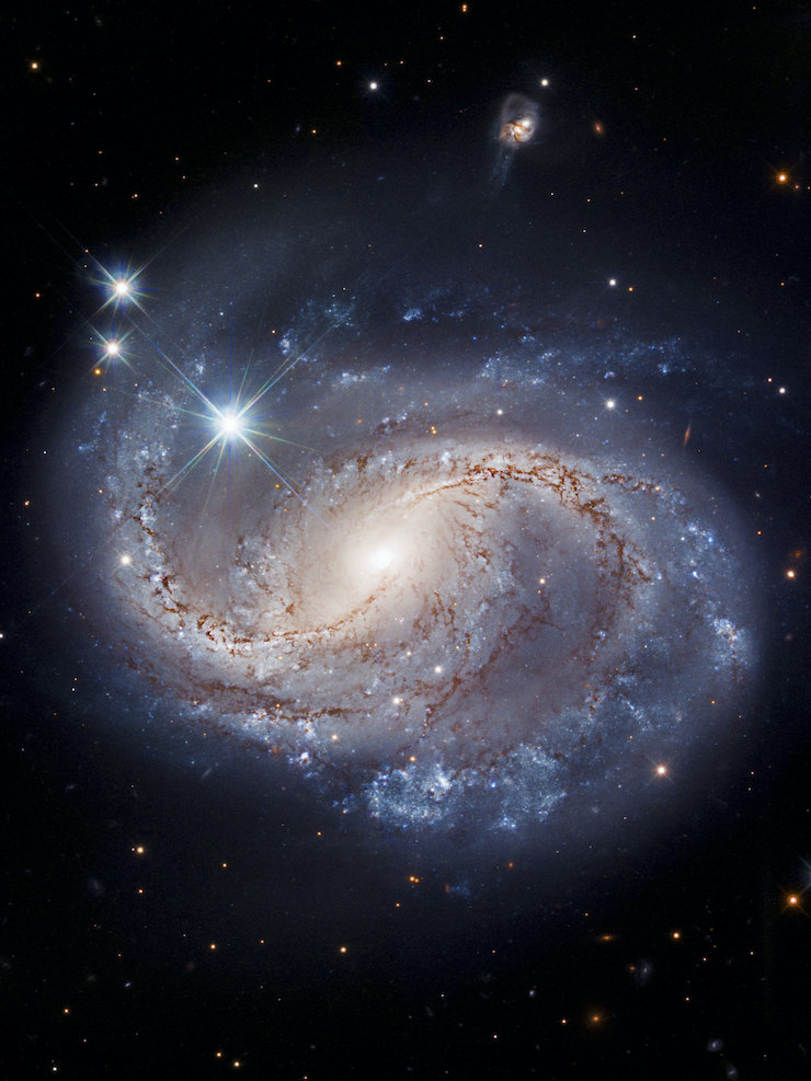 NGC 6956 is a barred spiral galaxy with a yellowish center and two primary arms spiraling away from the barred middle. The background is deep black with isolated stars and a few small galaxies. In the middle third, the arms are yellowish with dark red, irregular streaks throughout. As the arms reach the outer third of the galaxy, they turn blue. Subscribe to Deep Space Opera Newsletter!
