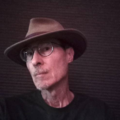 Portrait of author Ray N. Franklin, brown felt hat, light brown skin tone, eyeglasses, black background with heavy shadows.