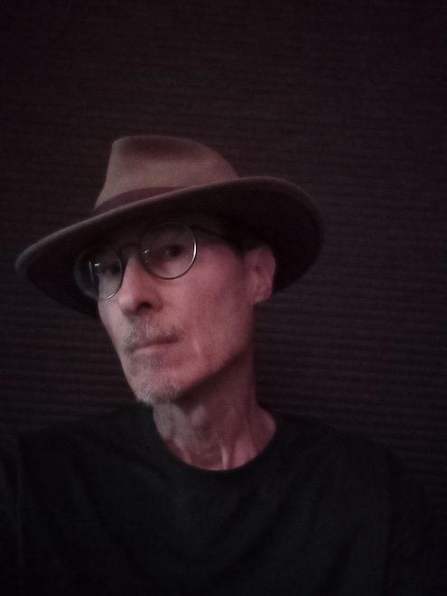 Portrait of author Ray N. Franklin, brown felt hat, light brown skin tone, eyeglasses, black background with heavy shadows.